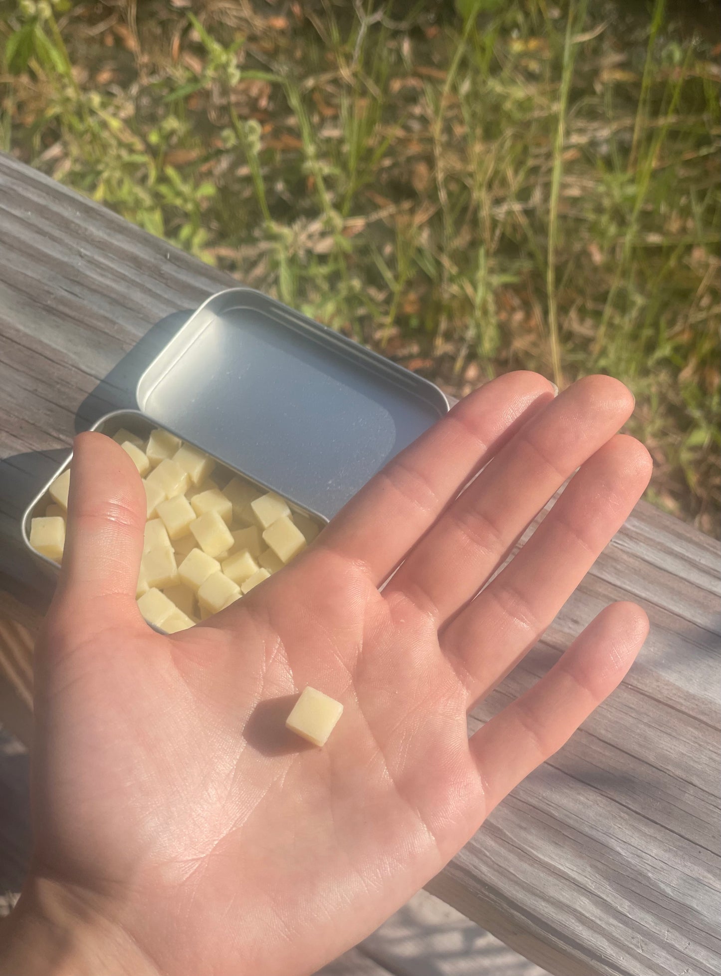 A single cube sits in the palm of a hand. The lotion cube is thinner and shorted than the width of the pinky. A full tin of lotion bars is in the background. The image is outside suggesting that the tin of lotion minis are excellent for travel and on the go