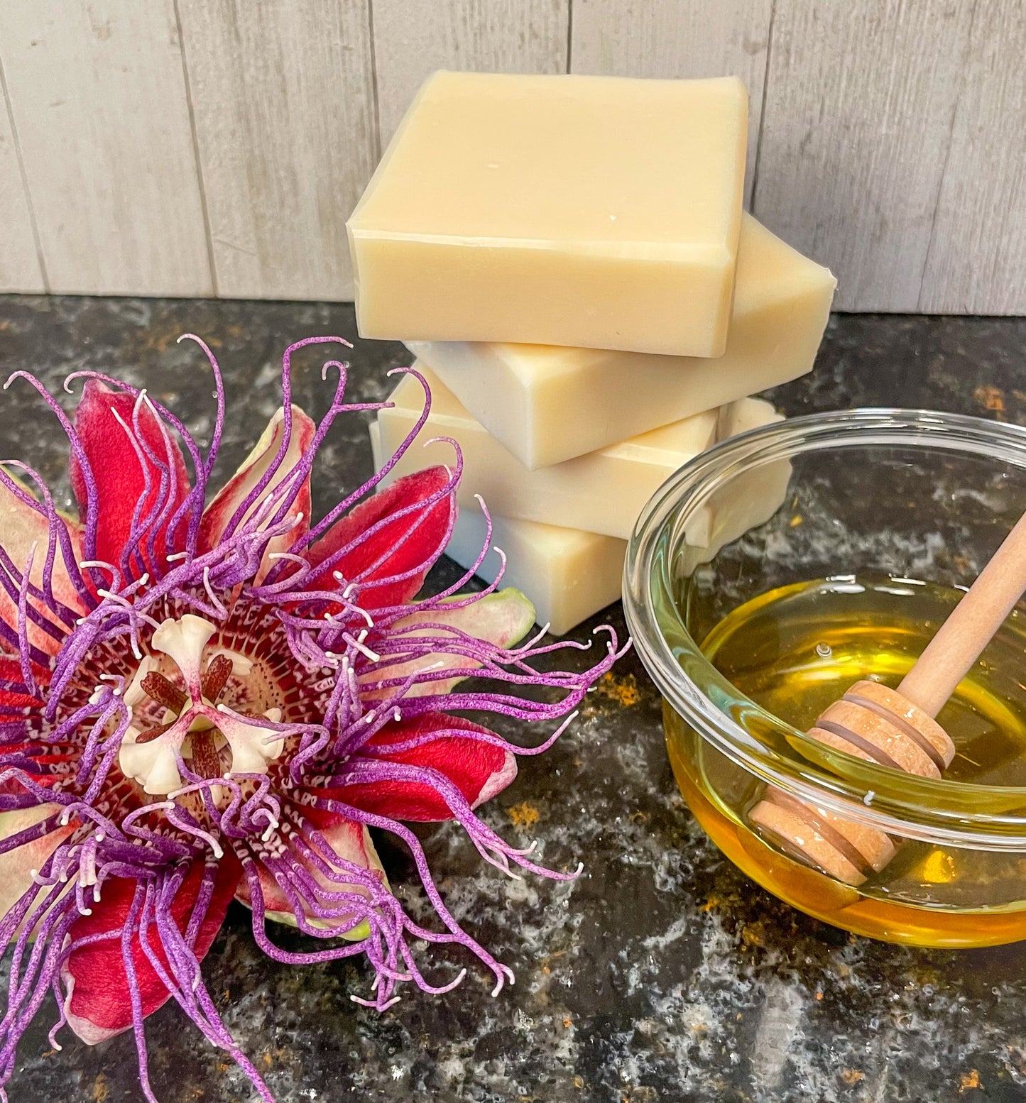 A stack of honey passionflower handmade soap stands next to a small pot of honey and a honey wand. A bright and in full bloom maypop purple passionflower sits to the left. The soap is naturally colored by the passionflower and honey