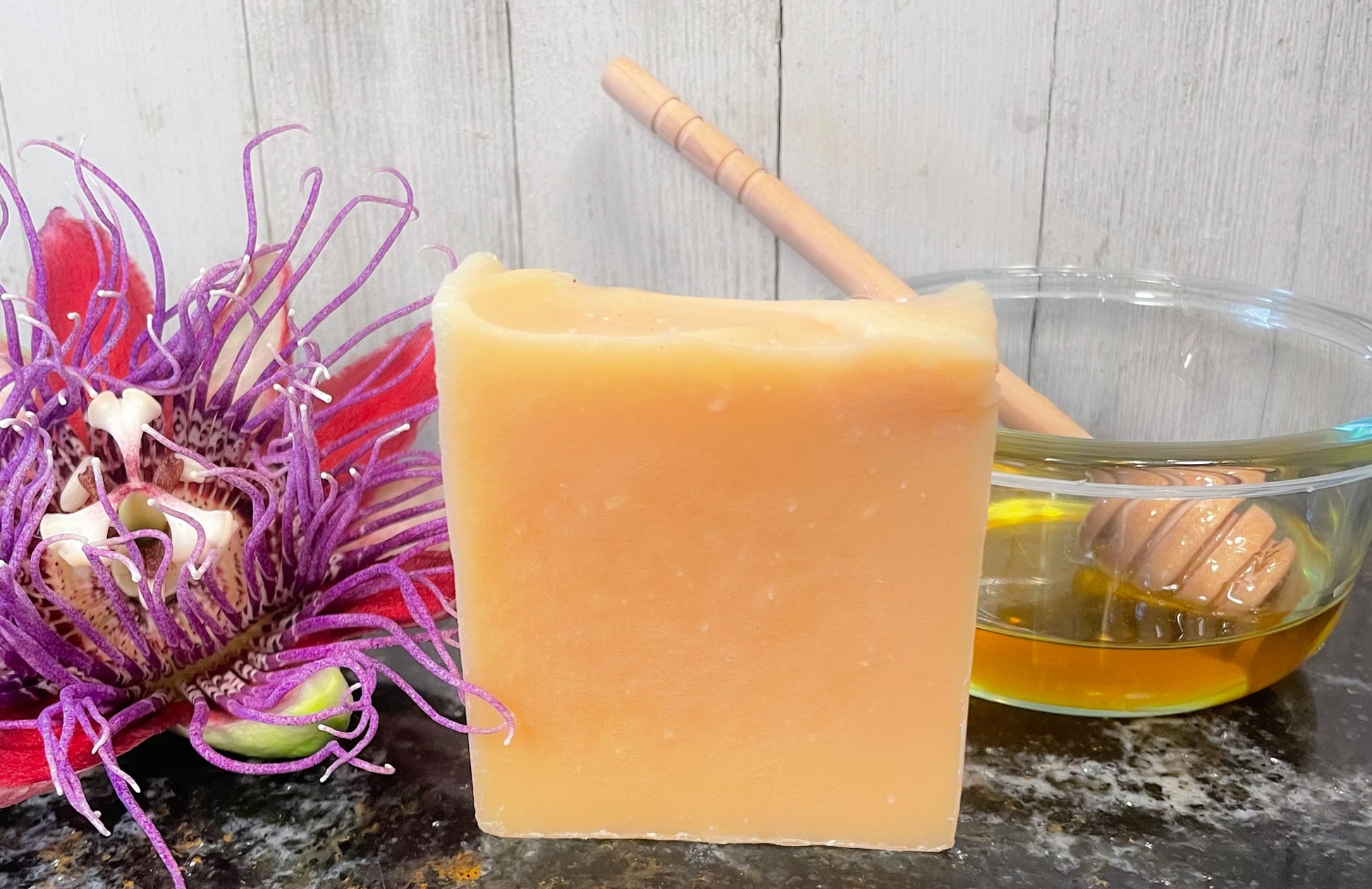 A bar of honey passionflower handmade soap stands next to a small pot of honey and a honey wand. A bright and in full bloom maypop purple passionflower sits to the left. The soap is a golden amber color which comes from the passionflower and honey