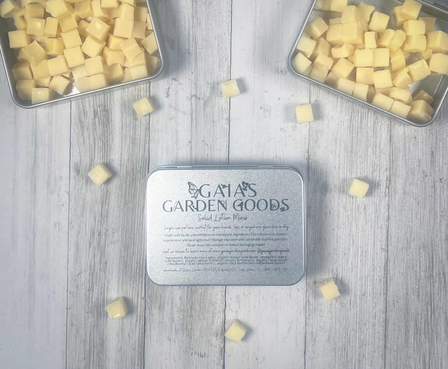 An open tin of Gaia's Garden Goods solid lotion minis is displayed against a light wooden background. The tin, filled with small, cube-shaped, yellow lotion bars, sits beside more scattered bars. The label on the tin provides product information, highlighting the organic and locally sourced ingredients