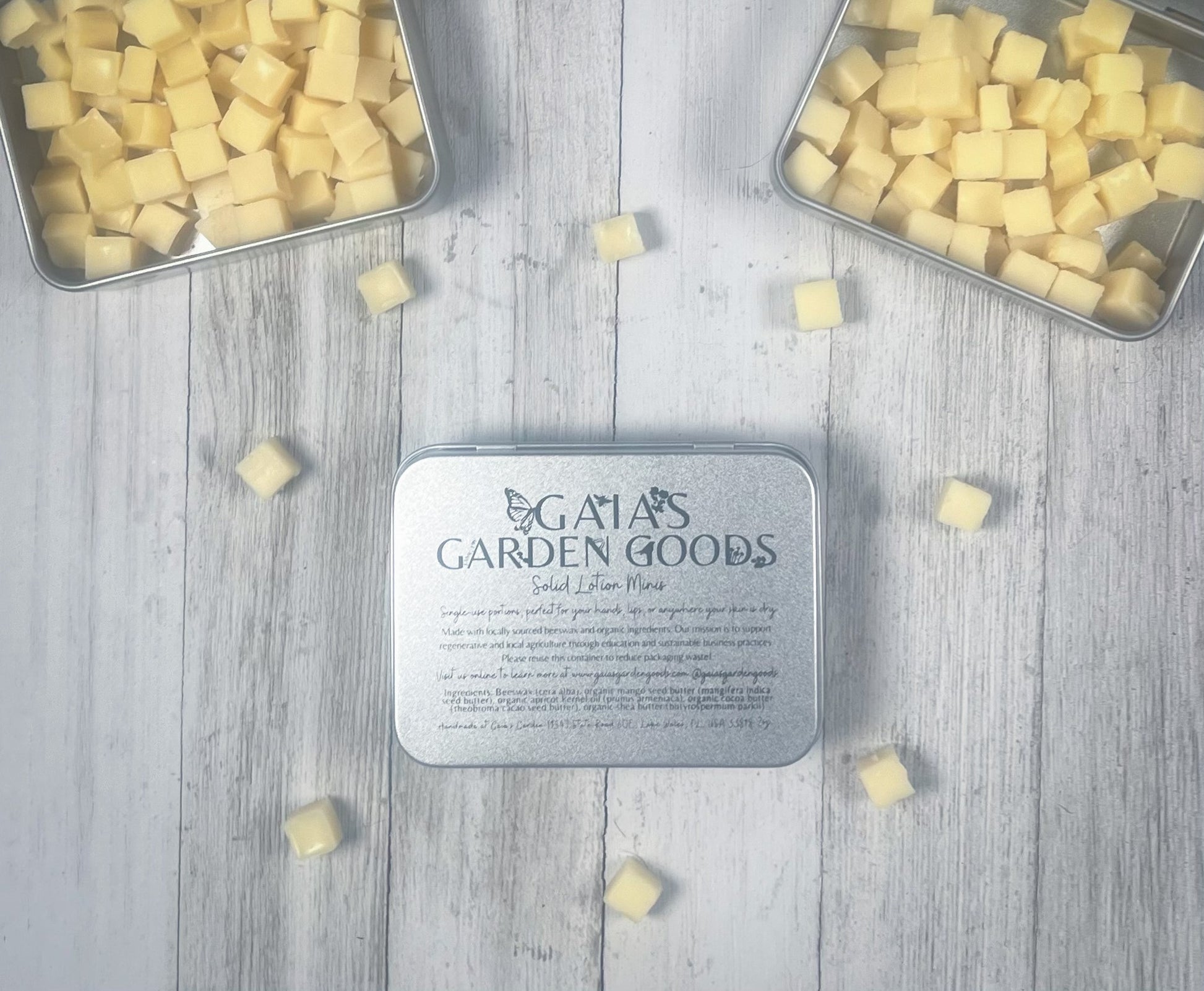 An open tin of Gaia's Garden Goods solid lotion minis is displayed against a light wooden background. The tin, filled with small, cube-shaped, yellow lotion bars, sits beside more scattered bars. The label on the tin provides product information, highlighting the organic and locally sourced ingredients