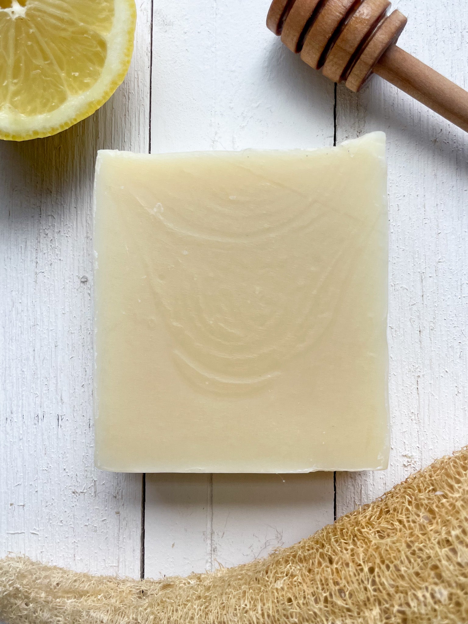 Honey lemonade citrus scented soap. surrounded by an organic loofah sponge, a lemon and a honey dipper. the soap is a pale off white color with subtle swirls rippling through the soap. the scene feels holistic and natural