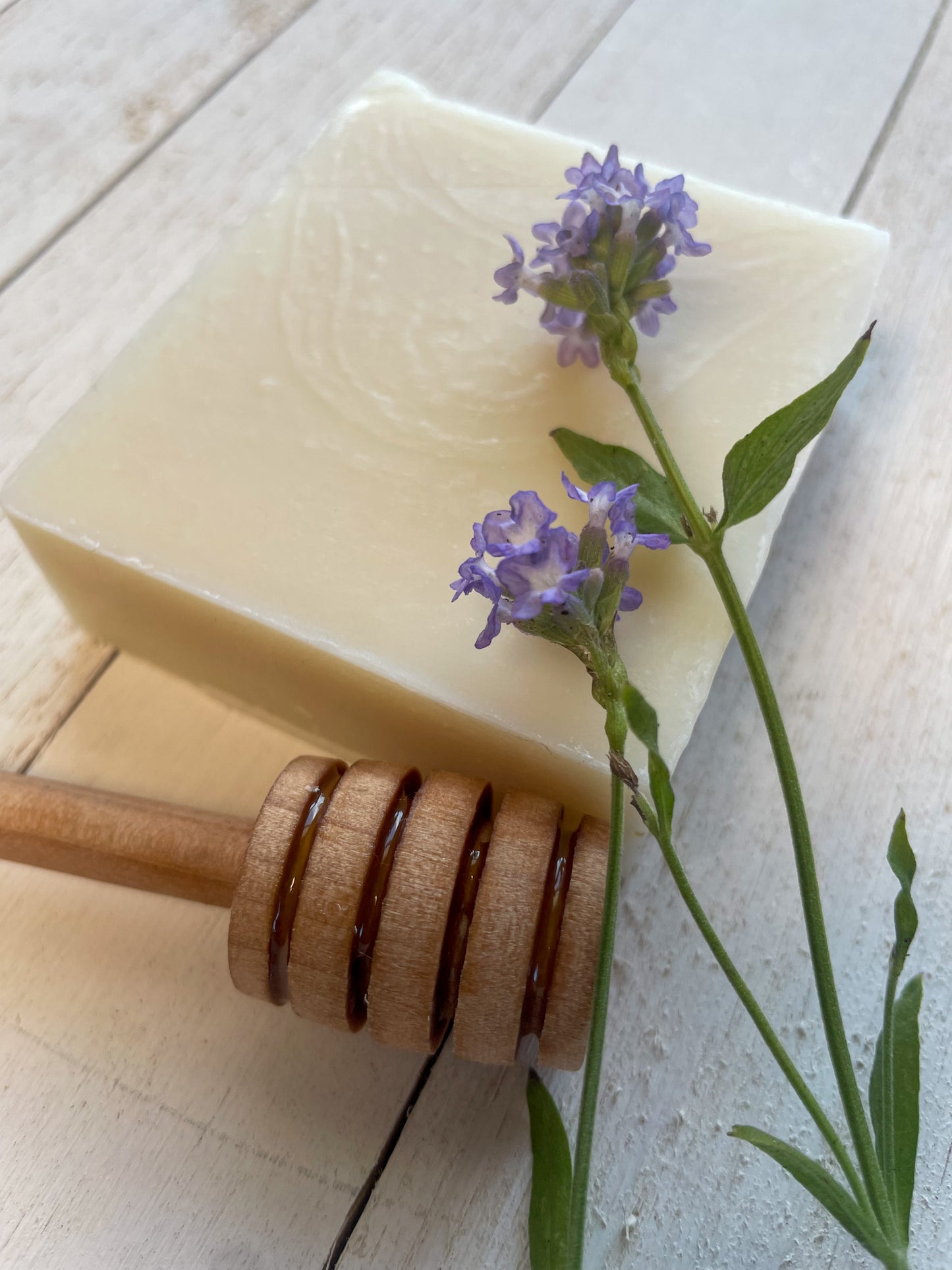 Honey Oatmeal Lavender Soap Bar lays flat against a white panel board. There are 2 springs of lavender and a honey stick laying besides the soap. The image is soothing and naturalist. 