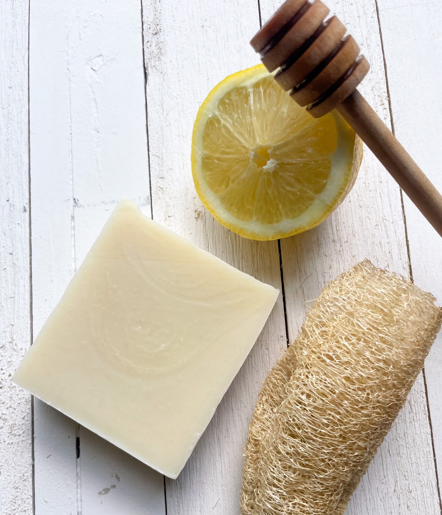 a bar of honey lemonade soap, a half a lemon, a honey wand, and a loofah sponge rest flat on a naturalistic looking wood panel board